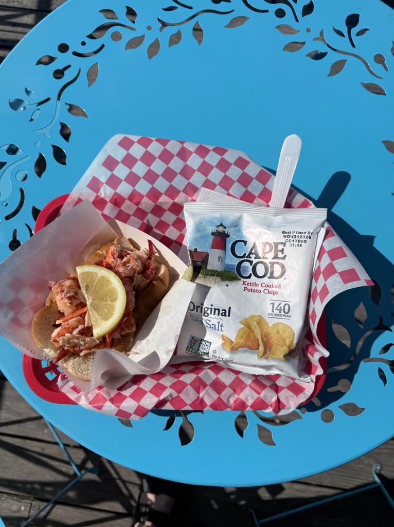 A lobster roll topped with a lemon wedge served on a checkered paper, accompanied by a bag of Cape Cod Original Kettle Cooked Potato Chips, on a blue decorative table.
