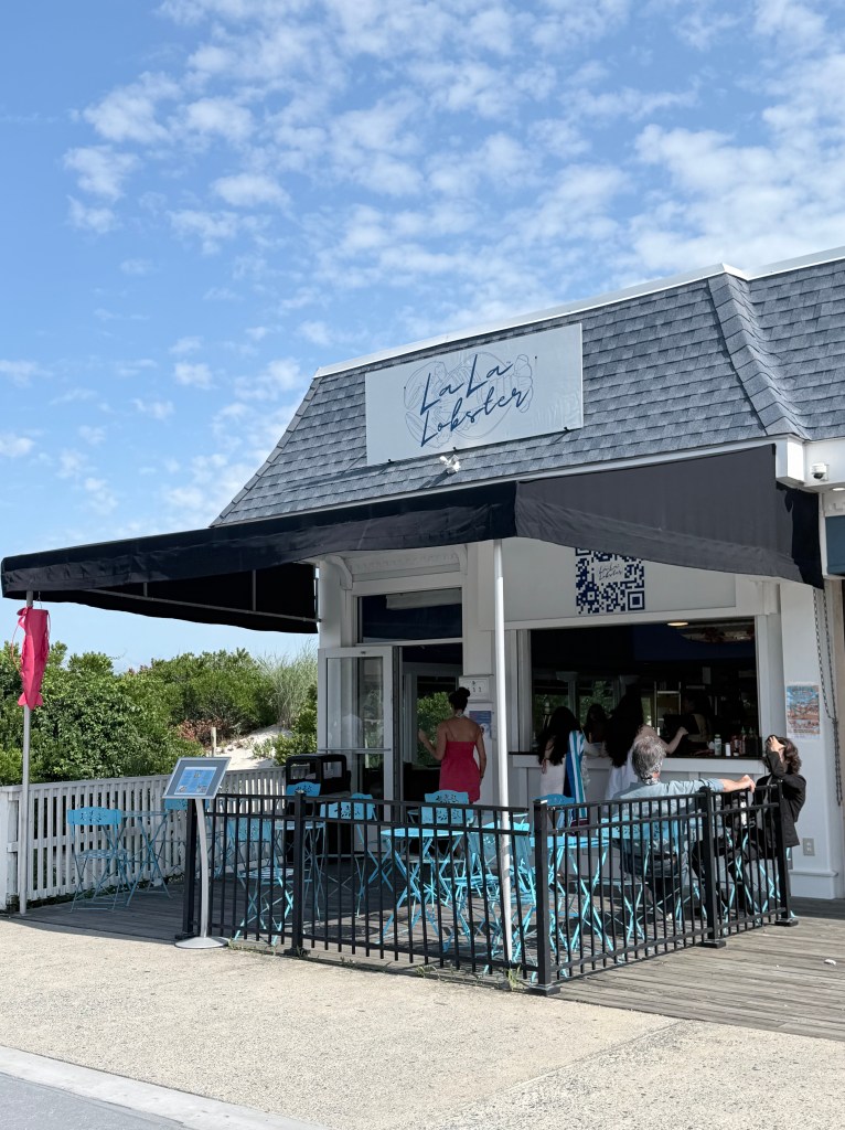 Exterior of La La Lobster restaurant featuring a black awning and turquoise chairs, with people dining and ordering at the counter on a sunny day.